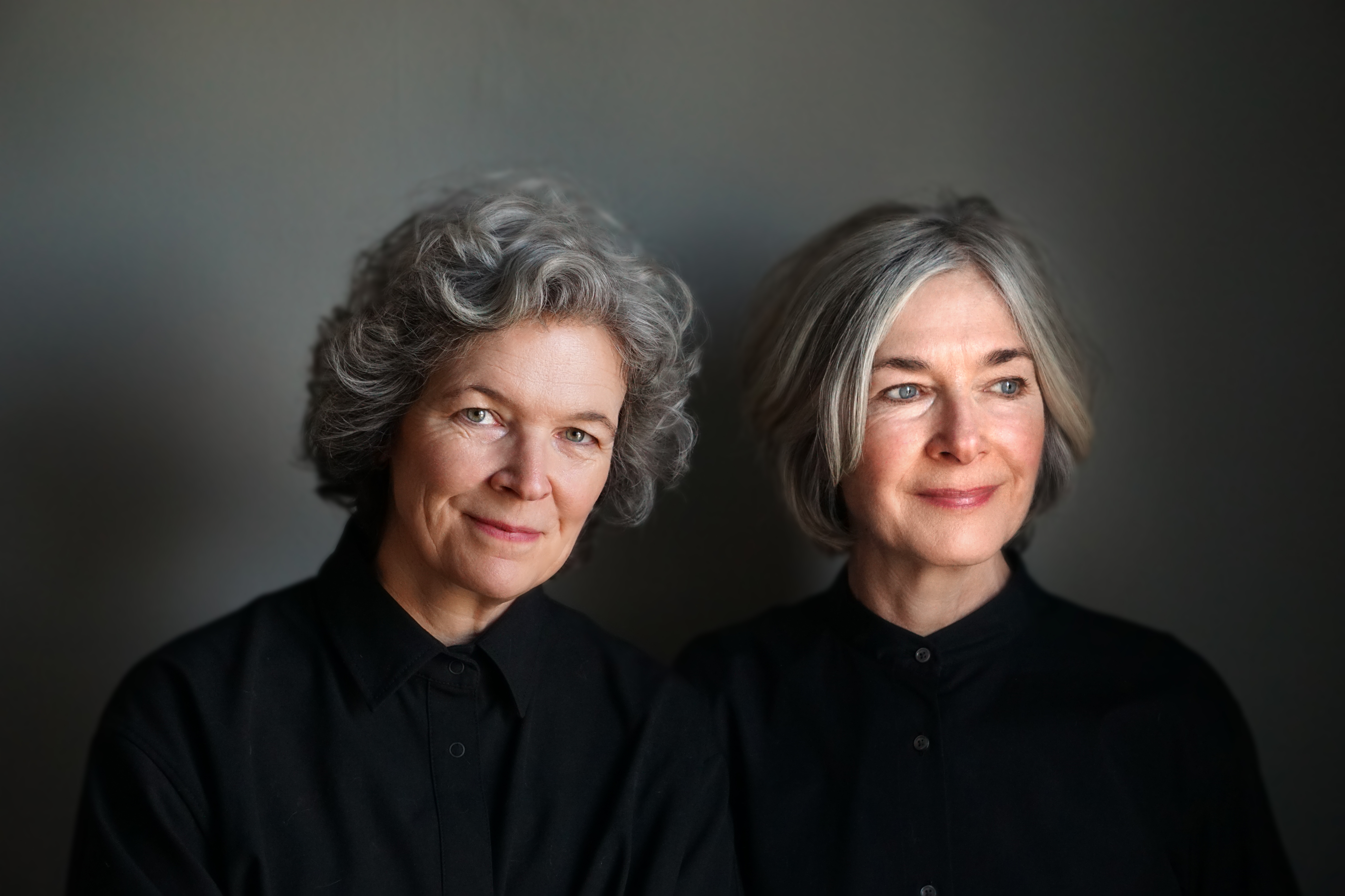 A portrait of Amanda Forbis and Wendy Tilby standing beside one another. Both are white women in their 60s with short grey hair. Amanda is on the left and is looking straight at the camera, Wendy is on the right and looks off into the distance.