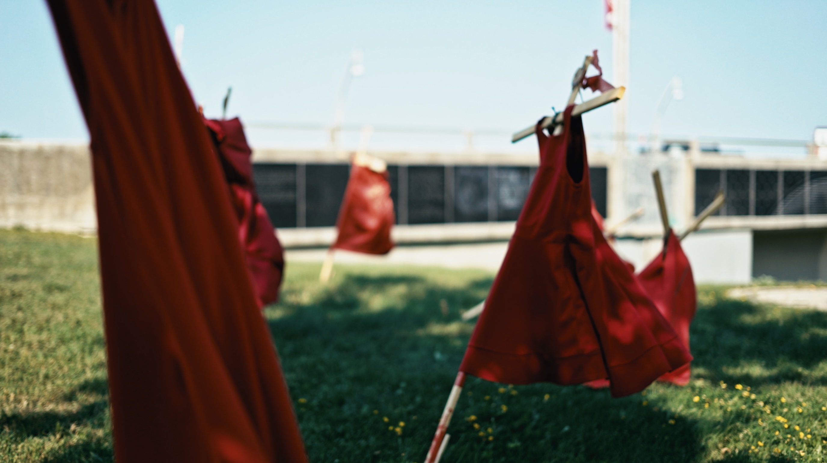 A still from Nova Weipert’s film #SearchTheLandfill. In the foreground, red dresses hang from tilting wooden crosses in a grassy field.